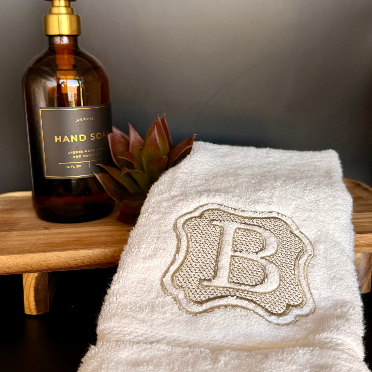 White towel personalized with a monogram 'B' next to a bottle of hand soap on a wooden surface.