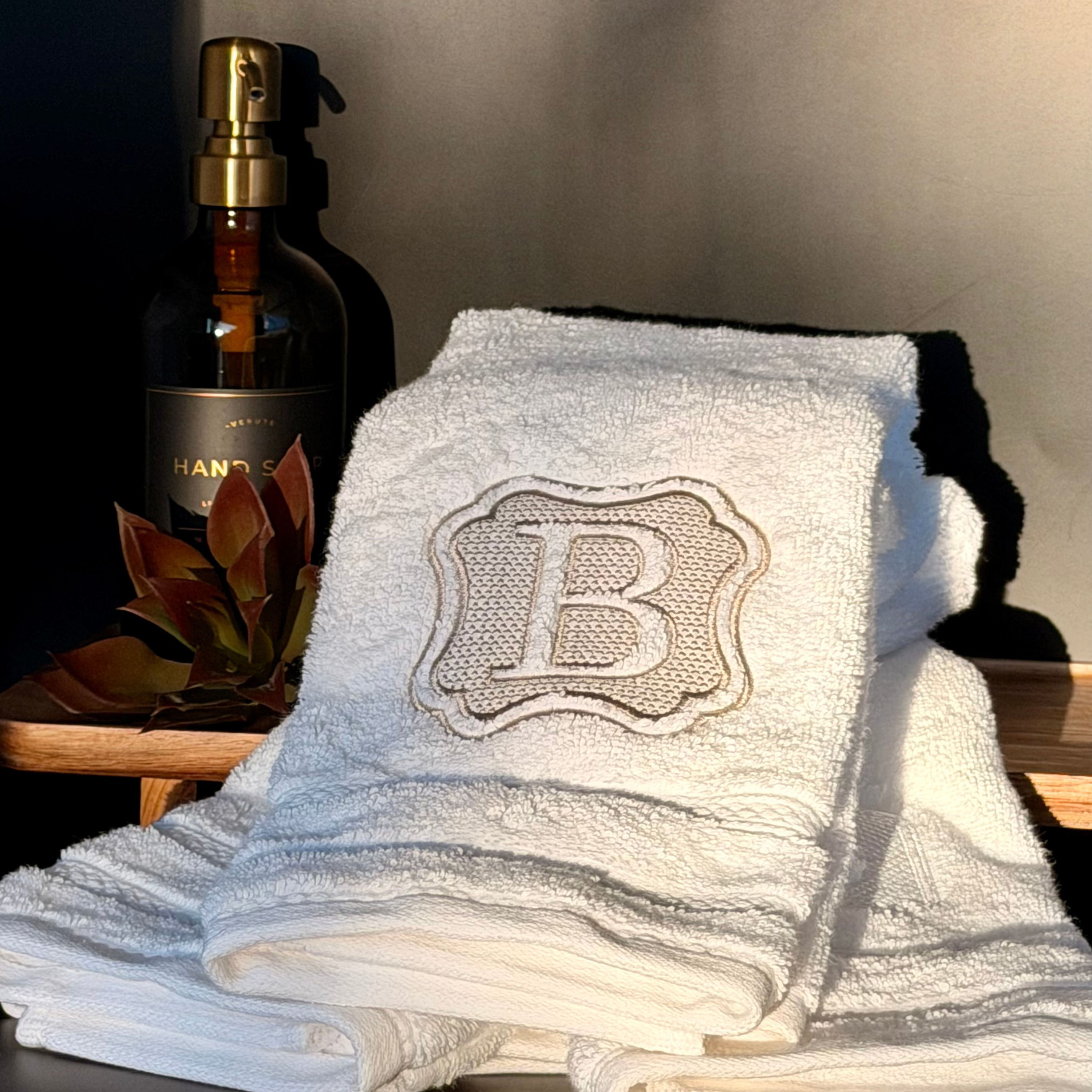 White luxury towel with a custom embroidered monogram next to a bottle of hand soap on a wooden surface.