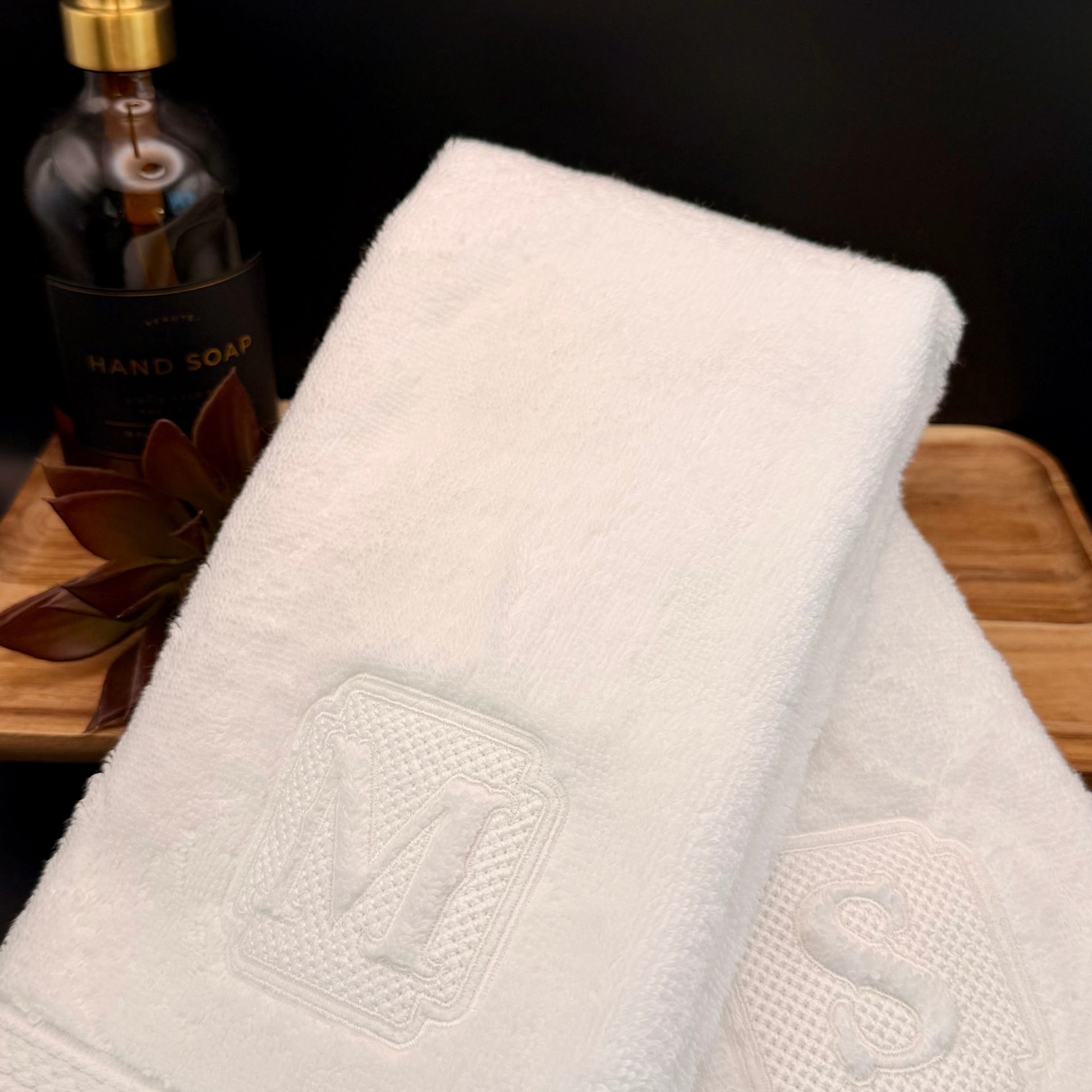 White luxury towel personalized with a monogram on a wooden surface next to a bottle of hand soap.