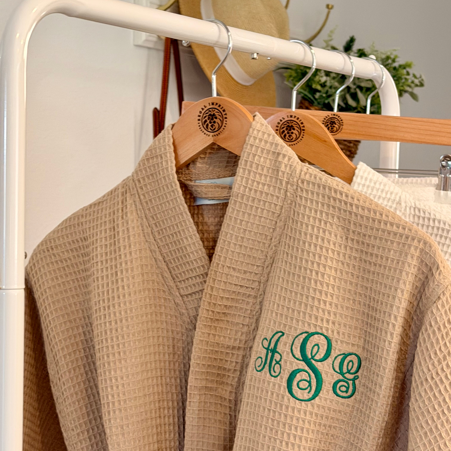 A beige waffle-textured robe hanging on a wooden hanger with a green embroidered monogram on the chest, displayed on a white clothing rack.